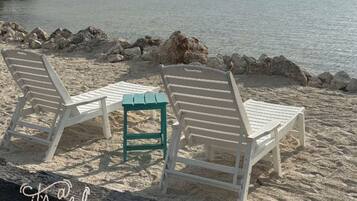 Une plage avec des chaises longues blanches et une table verte, un rivage rocheux et une mer calme sous un ciel nuageux.