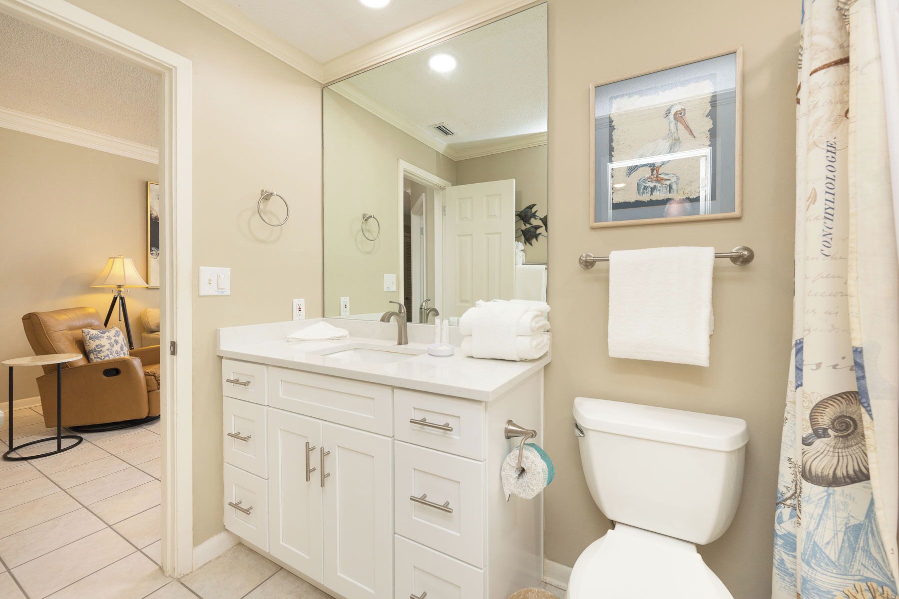 A bathroom with a toilet, sink, and mirror. There is a towel rack with towels, a framed picture on the wall, and a small table with a lamp.