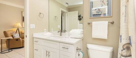 A bathroom with a toilet, sink, and mirror. There is a towel rack with towels, a framed picture on the wall, and a small table with a lamp.
