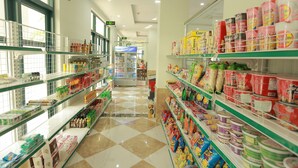 Um corredor de supermercado com prateleiras repletas de diversos produtos, incluindo bebidas, lanches e artigos para o lar.