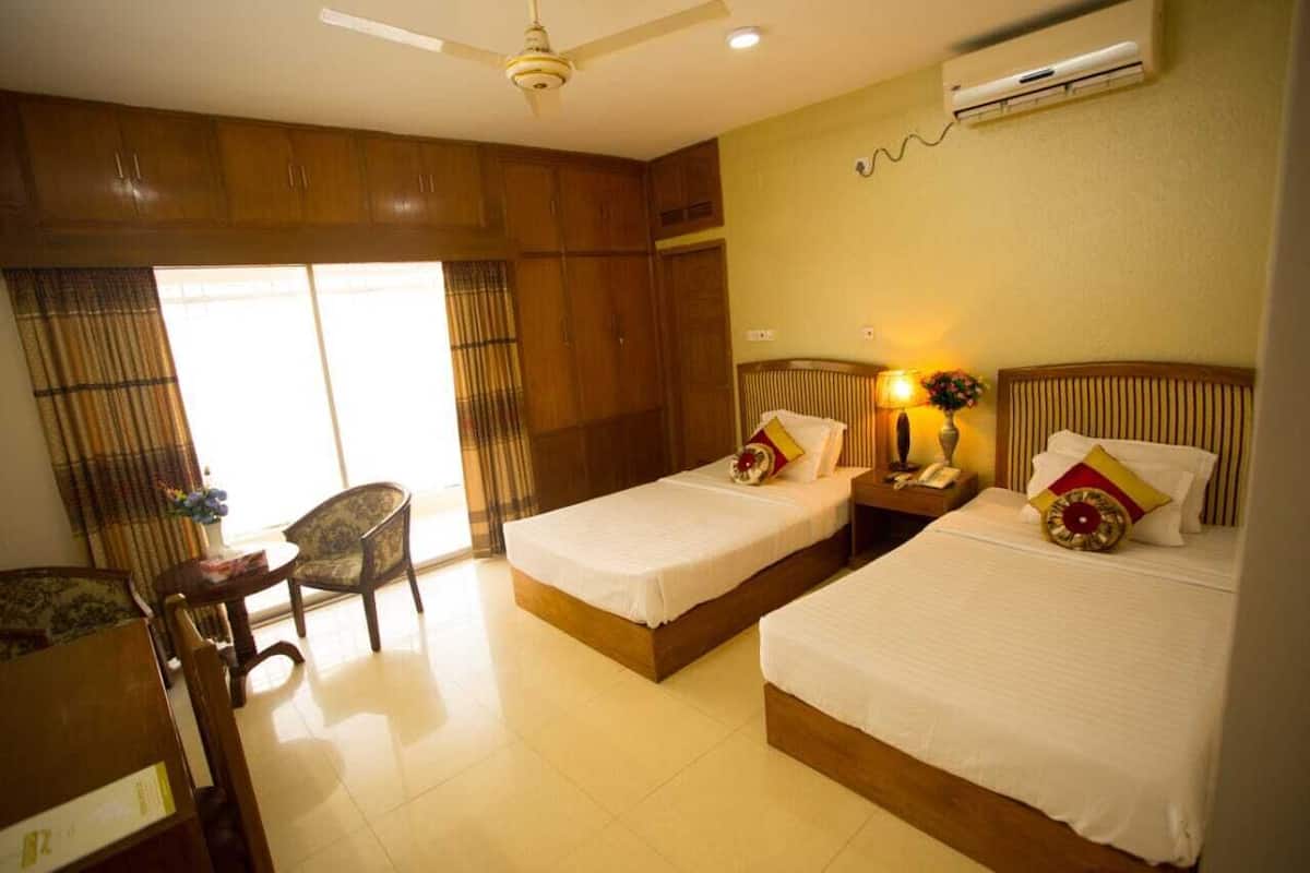 A hotel room with two beds, a desk, a chair, a ceiling fan, and an air conditioning unit.