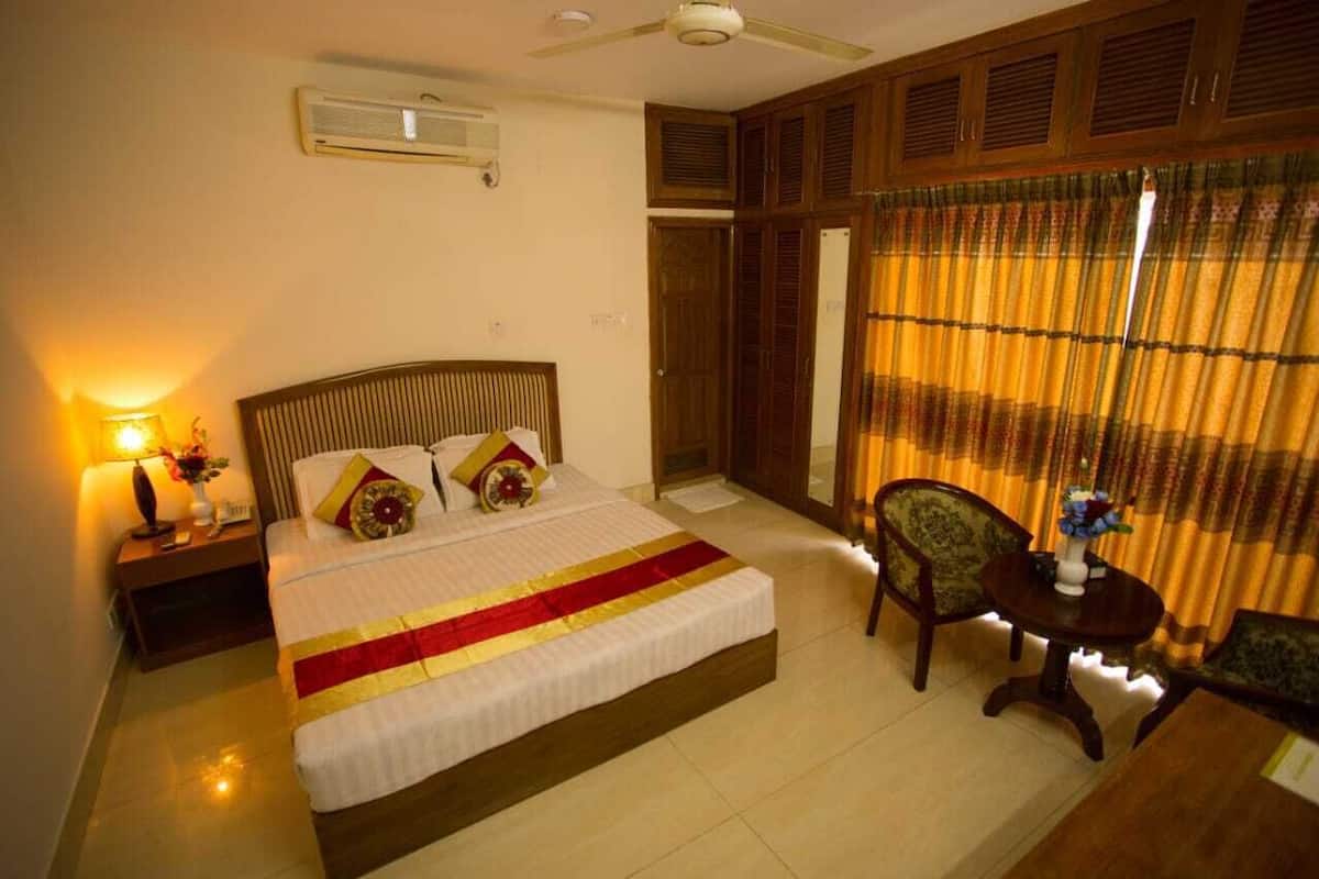 A hotel room with a bed, bedside table with a lamp, a chair, and a small table with a vase.