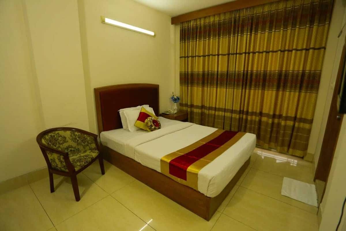 A hotel room with a bed, a chair, a bedside table with a lamp, and a window with curtains.