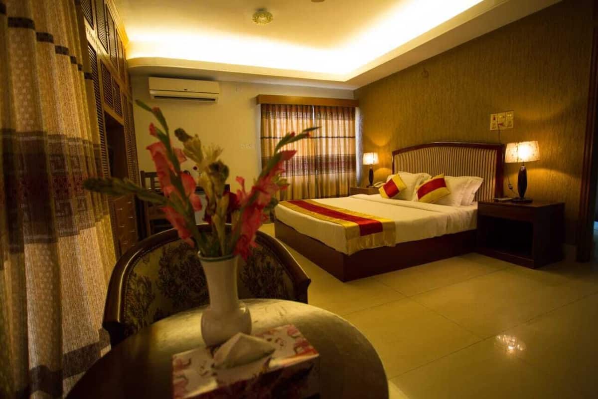 A hotel room with a bed, a bedside table with a lamp, a chair, and a vase with flowers.