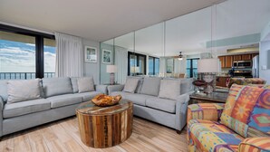 Een moderne woonkamer met een grijze bank, een kleurrijke fauteuil, een houten salontafel en een keuken op de achtergrond.
