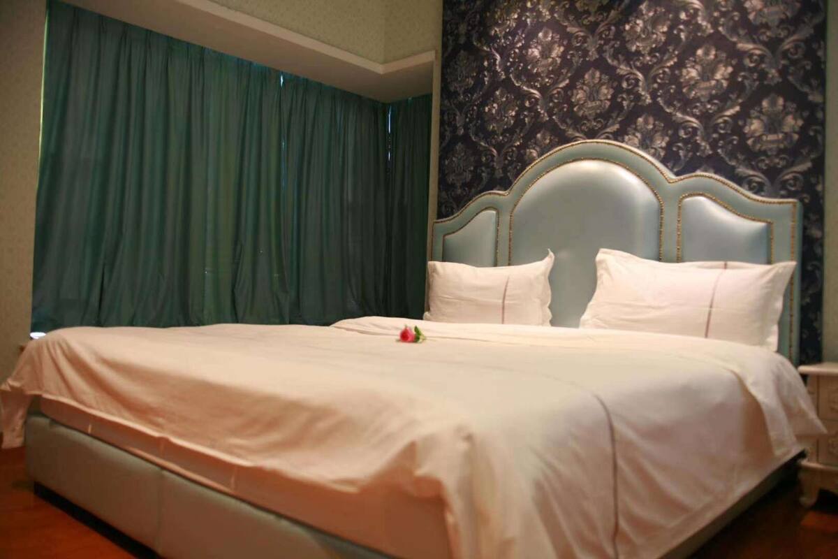 A neatly made bed with white linens and pillows, a blue headboard, and a dark patterned wall behind it.
