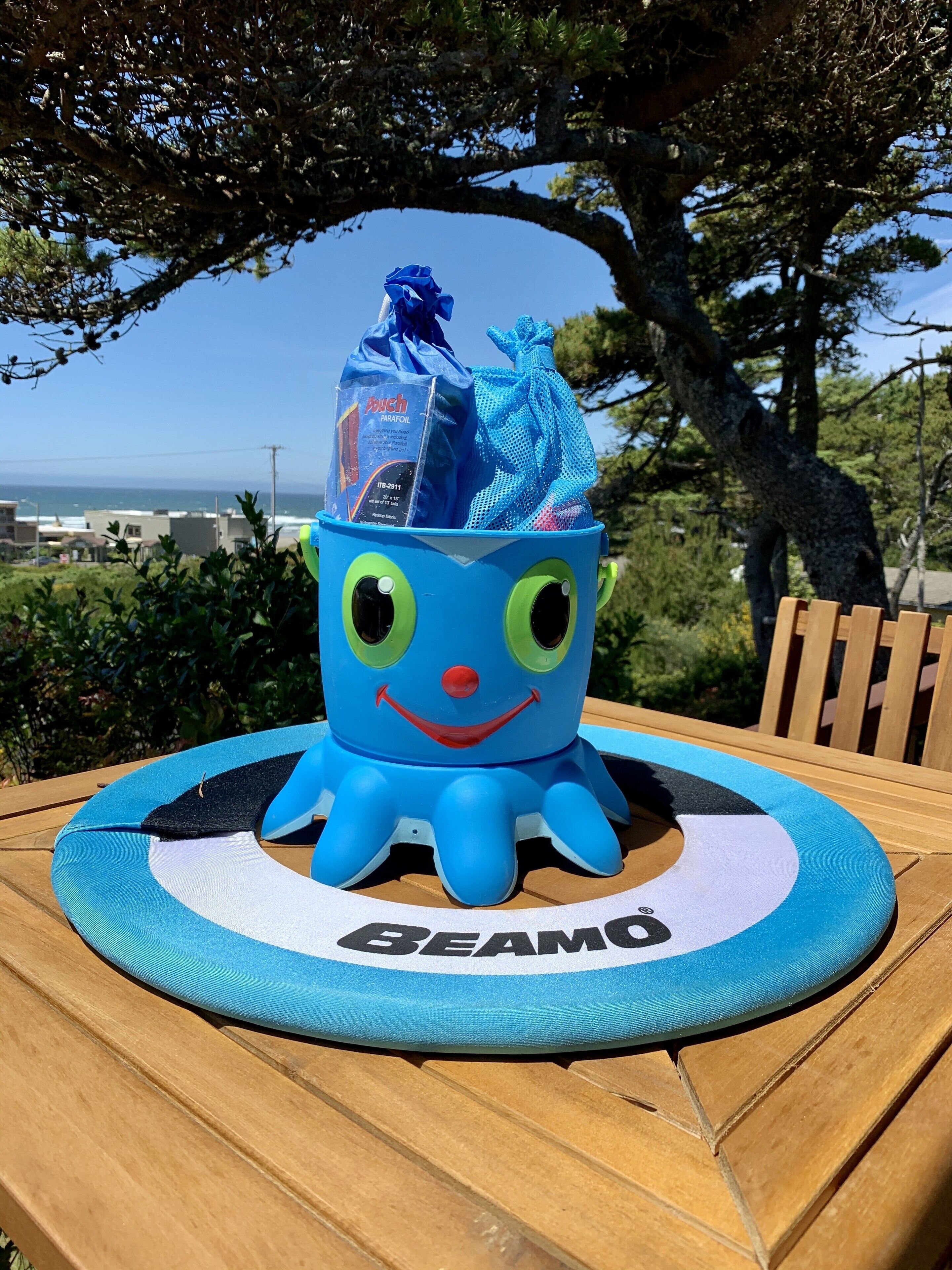 A blue inflatable pool float with a smiling face and the word "BEAMO" on a wooden deck with a view of the ocean and trees in the background.