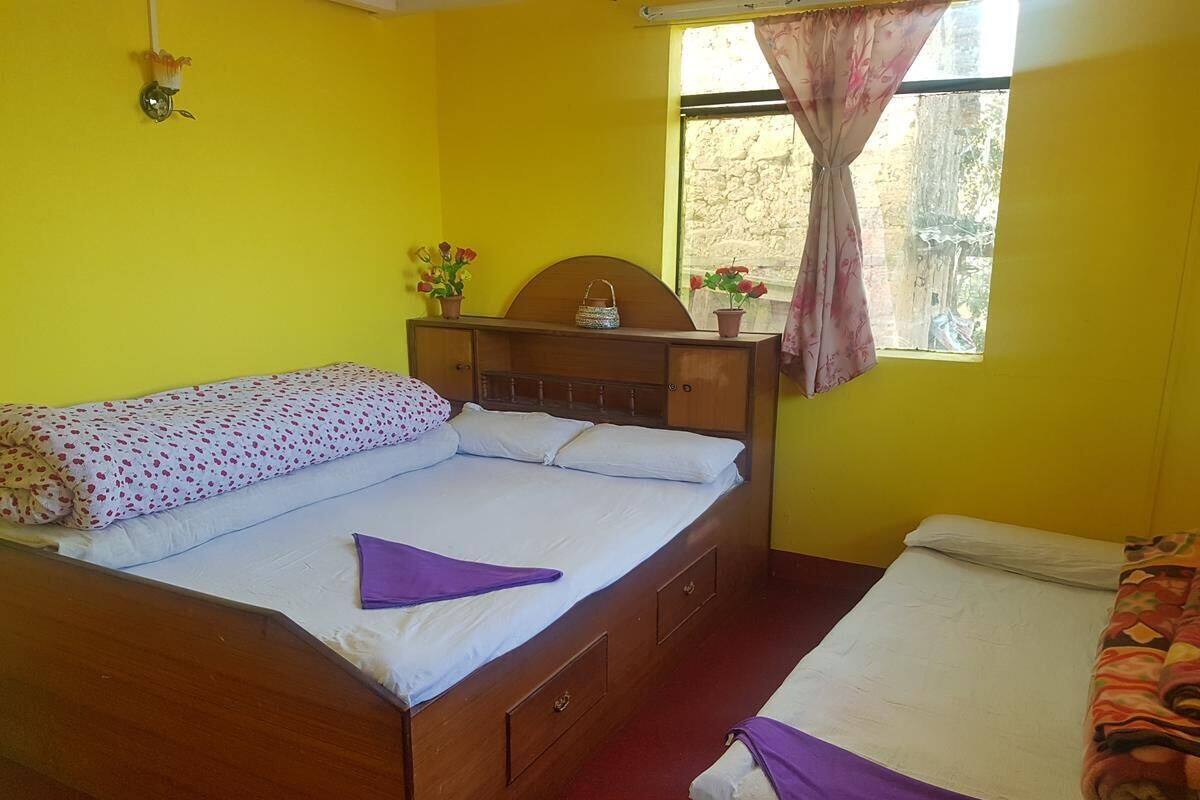 Una habitación con una cama de madera, una cama más pequeña, una ventana con cortinas y una lámpara fijada a la pared.