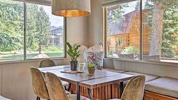 A dining area with a wooden table and chairs, a bench with cushions, and a view of a wooden cabin through large windows.