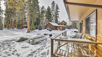 A wooden cabin with a deck, surrounded by snow-covered ground and tall pine trees.