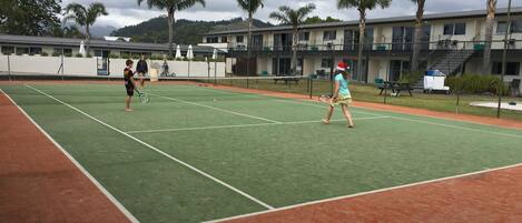 An outdoor tennis court with a net, surrounded by palm trees and a building in the background.