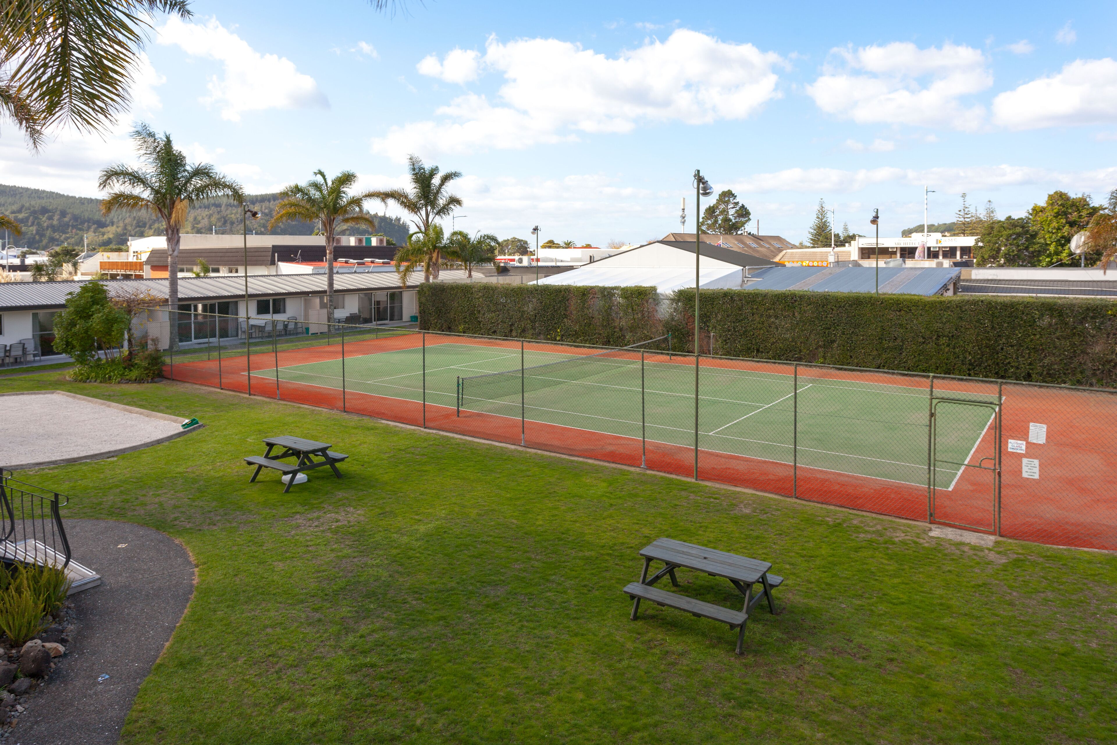An outdoor tennis court with picnic tables, surrounded by a fence and greenery.