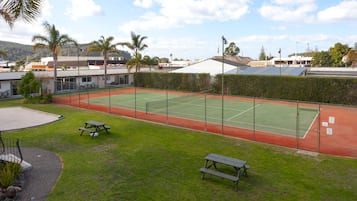An outdoor tennis court with picnic tables, surrounded by a fence and greenery.