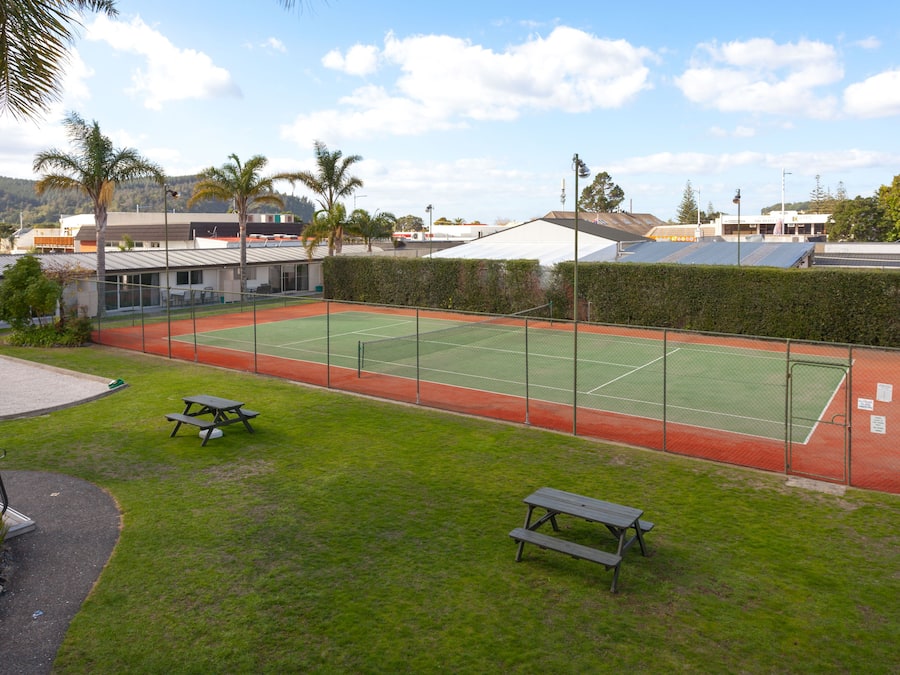 An outdoor tennis court with picnic tables, surrounded by a fence and greenery.