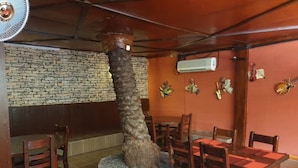 Ein Restaurantinneres mit einer Steinwand, Holzsesseln, einer zentral platzierten Baumskulptur und einer Klimaanlage.