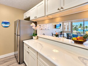 A modern kitchen with a stainless steel refrigerator, white cabinets, a marble countertop, and a view of the ocean through large windows.