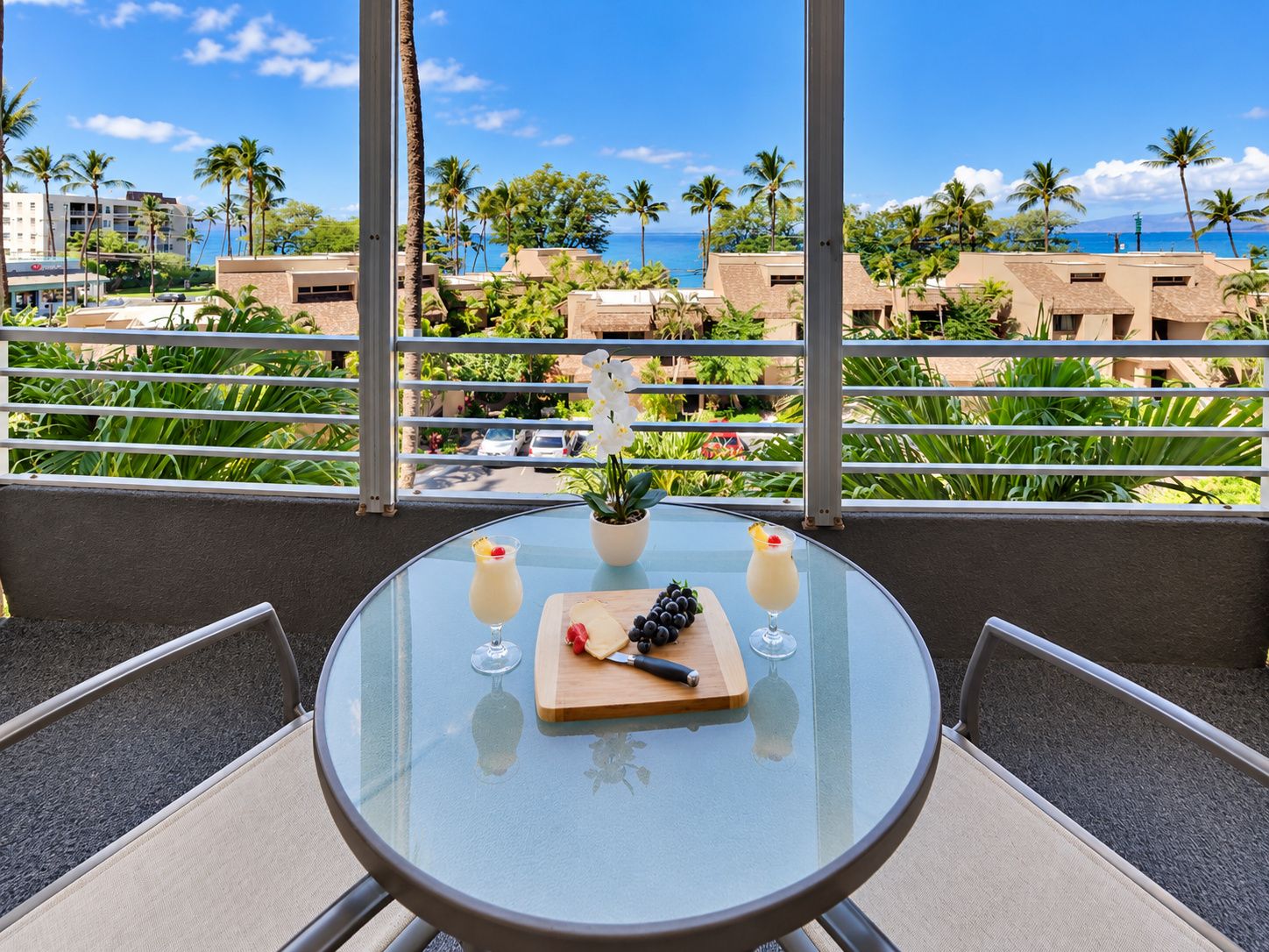 A round glass table with a small potted plant, two drinks, and a wooden board with fruits and a knife on a balcony overlooking a beach with palm trees and buildings.