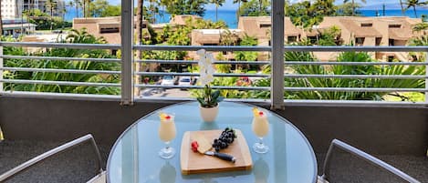A round glass table with a small potted plant, two drinks, and a wooden board with fruits and a knife on a balcony overlooking a beach with palm trees and buildings.