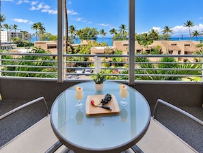 A round glass table with a small potted plant, two drinks, and a wooden board with fruits and a knife on a balcony overlooking a beach with palm trees and buildings.
