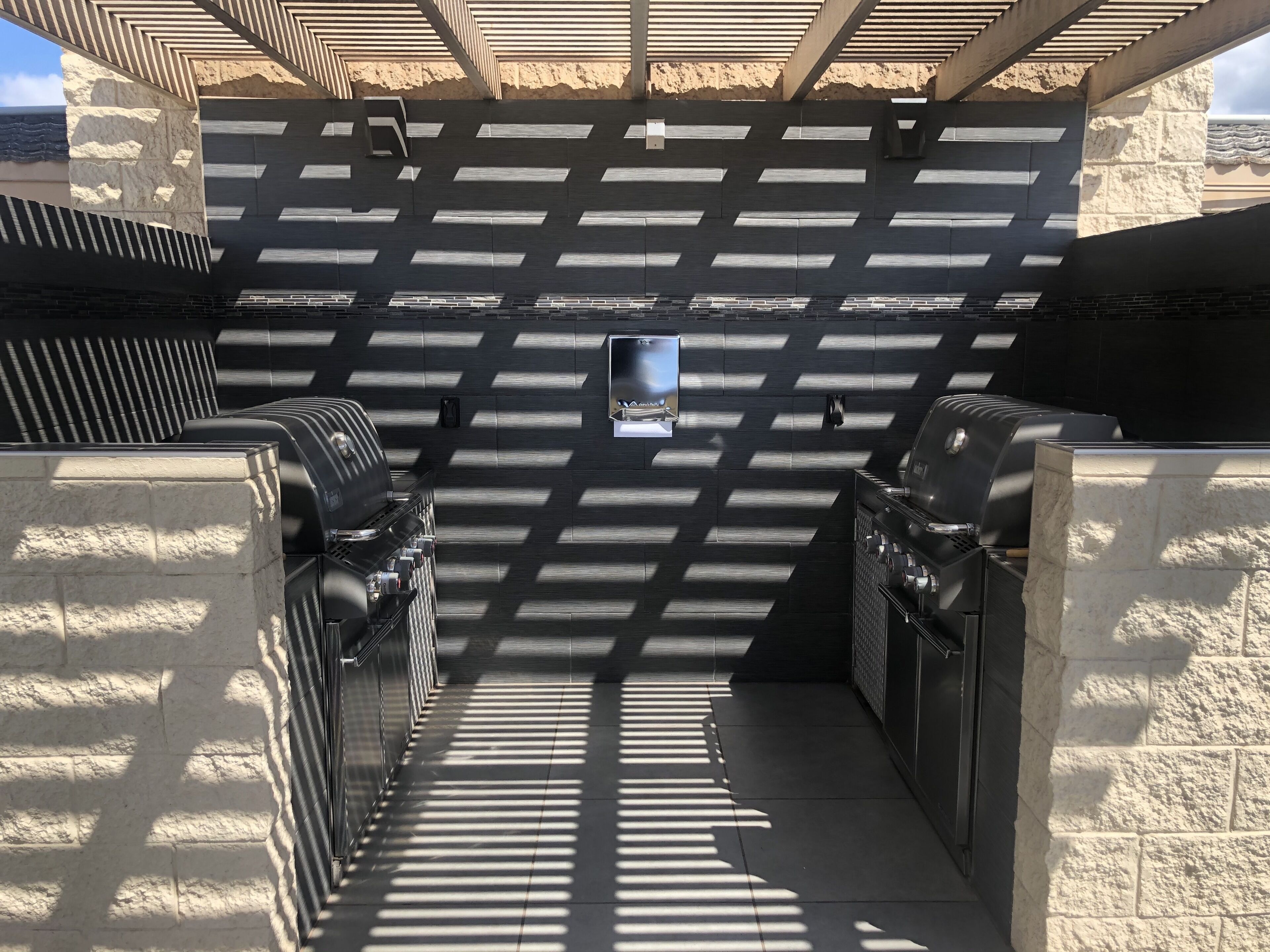 Een terras buiten met een ingebouwde barbecueplaats, stenen muren en een pergola voor schaduw.