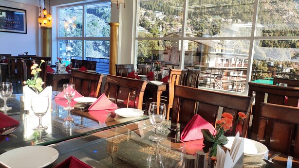 A restaurant with a glass table set for dining, wooden chairs, and a view of a mountainous landscape through large windows. - Hotel Shivani International (Dharamshala)