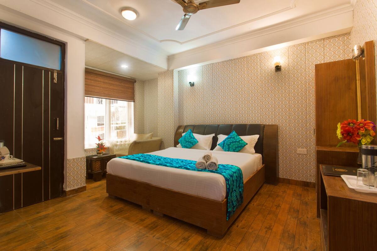 A hotel room with a large bed, wooden flooring, a desk with a laptop, a wardrobe, and a window with curtains.