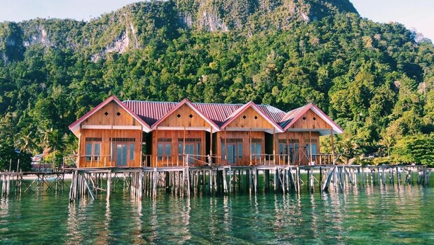 A row of wooden bungalows with red roofs, built on stilts over clear water, with a forested mountain in the background. - Ora Sunrise View Resort (Salemang)