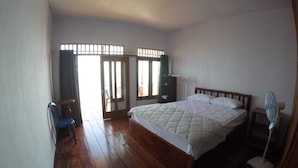 A bedroom with a wooden floor, a bed with a quilted blanket, a standing fan, and a window with bars. - Ora Sunrise View Resort (Salemang)