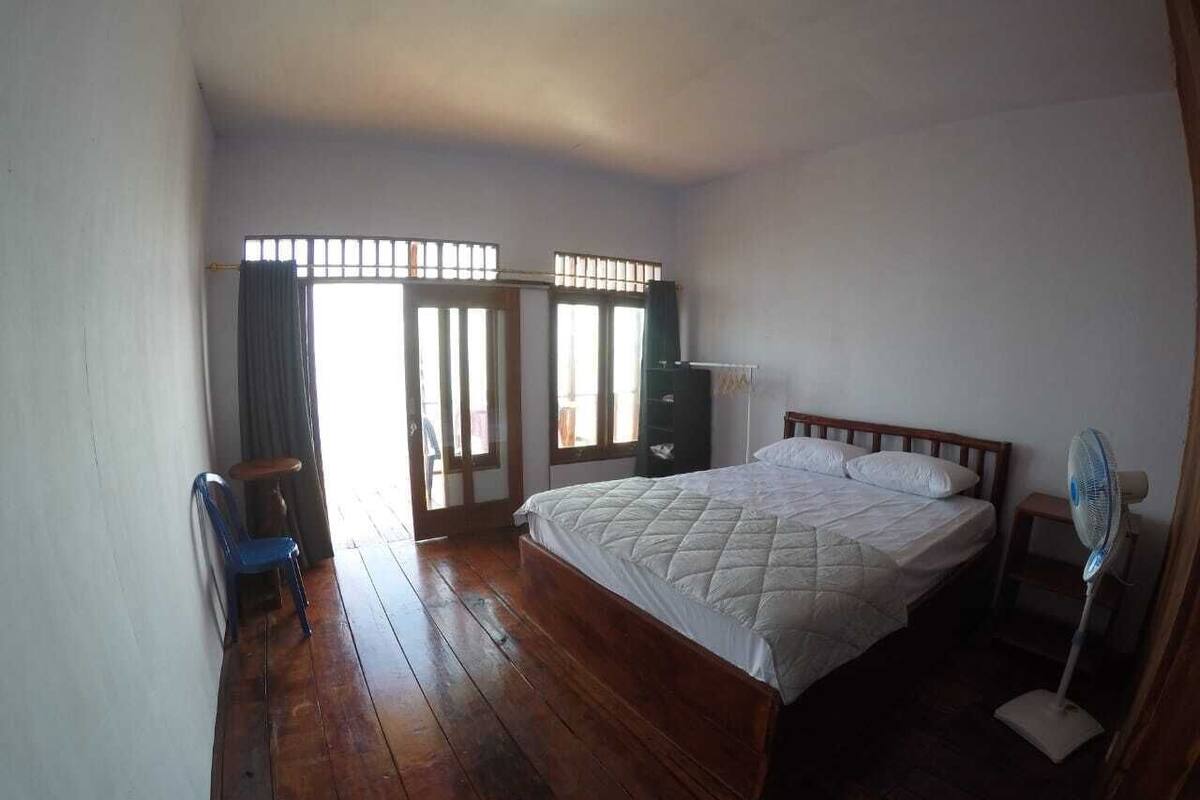 Une chambre avec un plancher en bois, un lit recouvert d’une couverture matelassée, un ventilateur sur pied et une fenêtre avec des barreaux.
