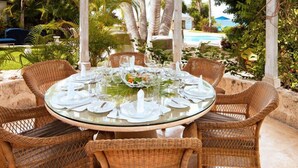 A round glass table set for six with wicker chairs, surrounded by lush greenery and a pool in the background.