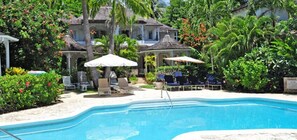 A pool area with lounge chairs and umbrellas, surrounded by lush greenery and a large house.