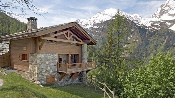Una cabaña de madera con base de piedra, un balcón y un paisaje montañoso.