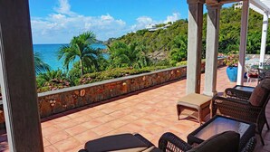 A covered outdoor area with wicker furniture, a stone wall, and a view of the ocean and hills. - Carnival Beach House Awaits: Relax in Style with Beachfront Views, A/C, and a Private Pool! (St John)