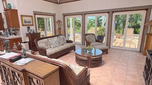 A spacious living area with a sofa, armchair, and coffee table, large sliding glass doors to a patio, and a wooden cabinet. - Carnival Beach House Awaits: Relax in Style with Beachfront Views, A/C, and a Private Pool! (St John)