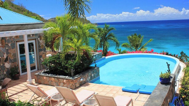 A pool with a view of the ocean, surrounded by palm trees and a stone building. - Carnival Beach House Awaits: Relax in Style with Beachfront Views, A/C, and a Private Pool! (St John)