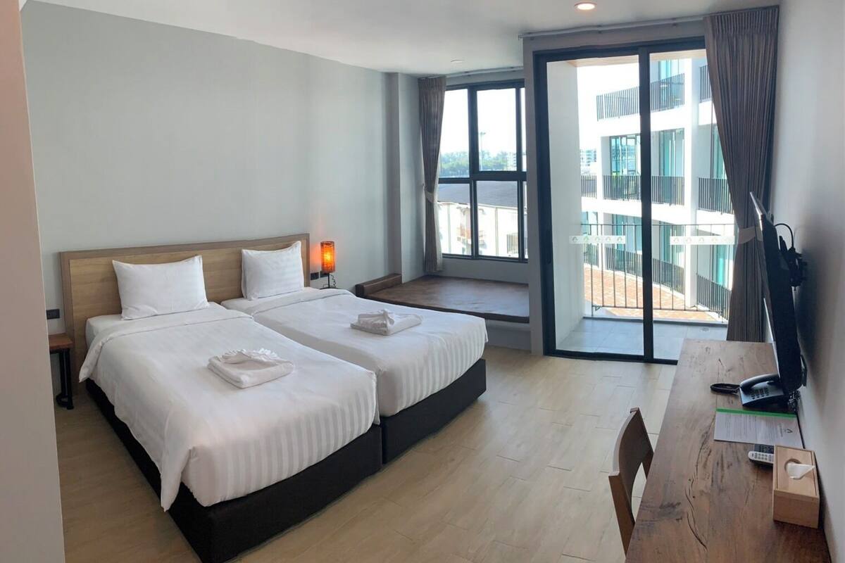 Habitación de hotel con dos camas, un escritorio, una silla, una ventana con vista a edificios y un balcón.
