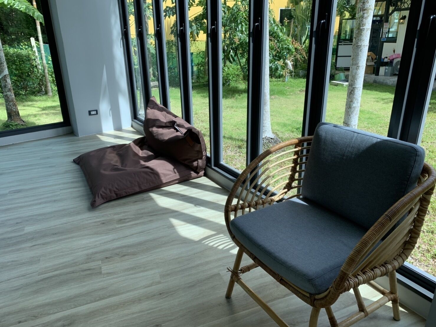 Una stanza con una poltrona in vimini e un pouf, vicino a grandi finestre che si affacciano su un giardino.