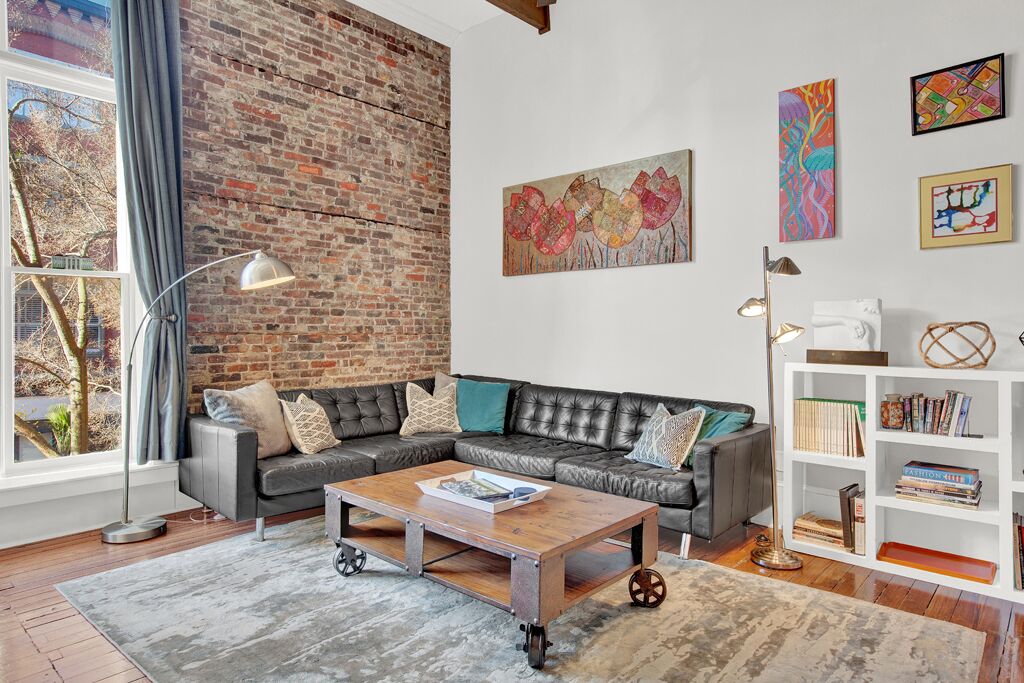 A modern living room with a brick wall, a sectional sofa, a coffee table with wheels, a bookshelf, and artwork on the walls.