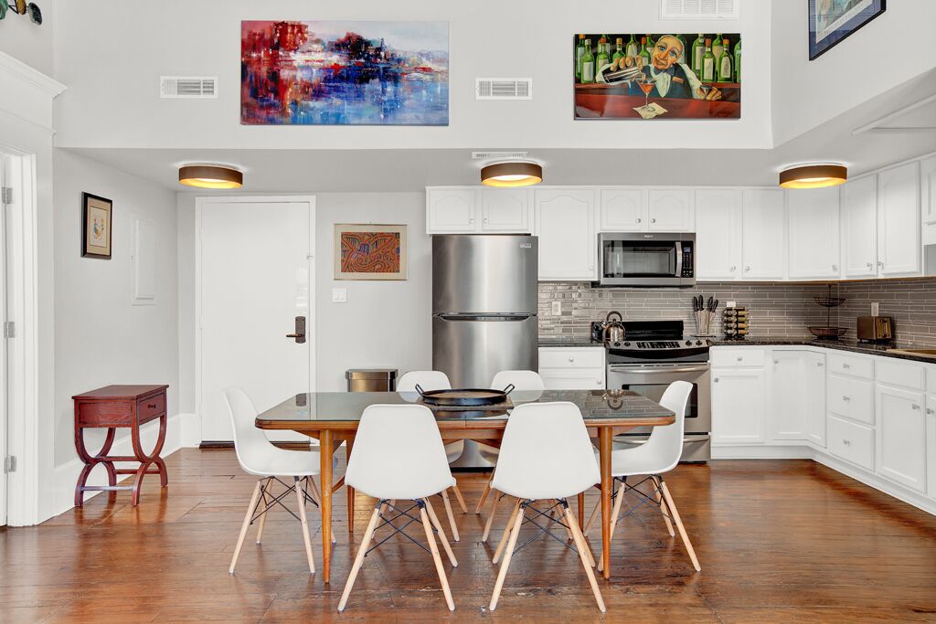 A modern kitchen with white cabinets, a stainless steel refrigerator, a microwave, and a gas stove. There are two white chairs around a wooden table.