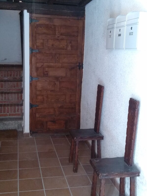 A room with a wooden door, two wooden chairs, tiled floor, and a white wall with electrical boxes.