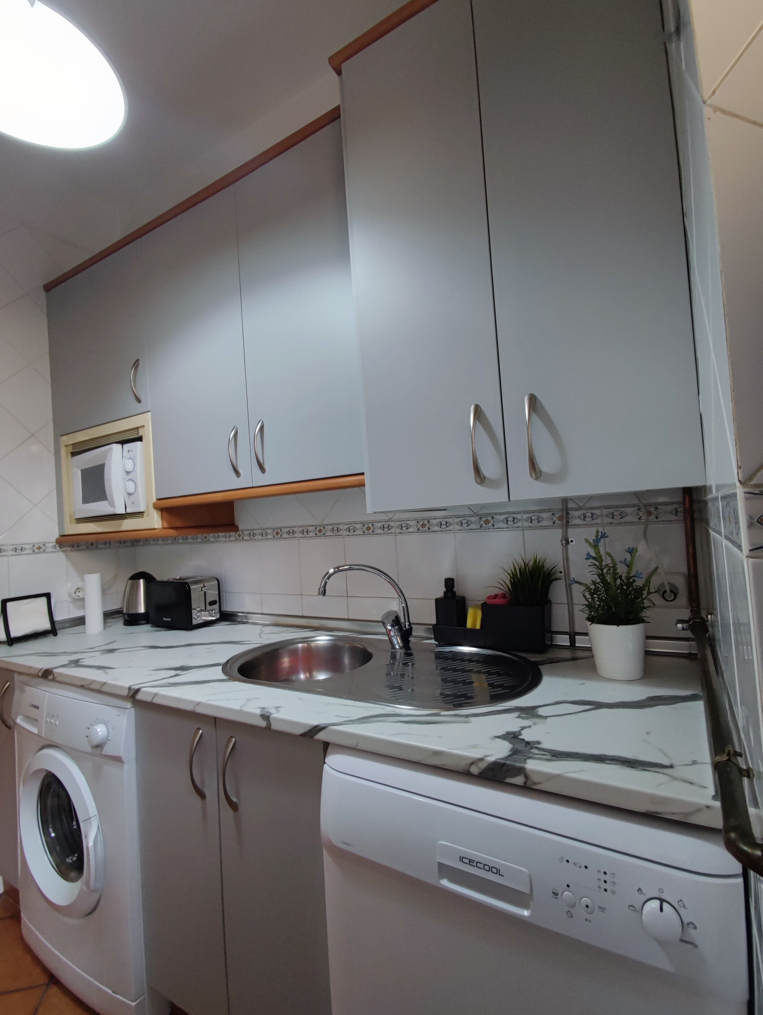 A modern kitchen with white cabinets, a built-in microwave, a stainless steel sink, a washing machine, and a dishwasher.