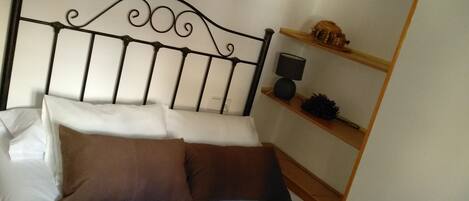 Una cama bien hecha con ropa de cama blanca y marrón, un cabecero metálico y un estante de pared con una lámpara y objetos decorativos.