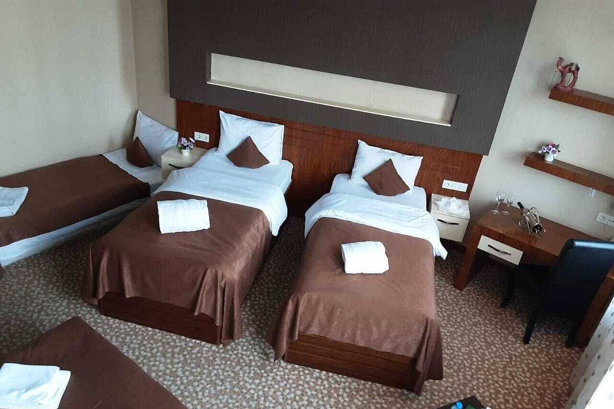 A hotel room with three single beds, a desk, a chair, and a small table with a bottle and glass.