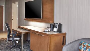 Quarto de hotel com uma mesa de madeira, televisão de tela plana, cafeteira e poltrona cinza com almofada estampada.