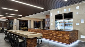 A modern bar area with a long counter, multiple stools, and a well-lit space with a wooden accent wall and a large window.
