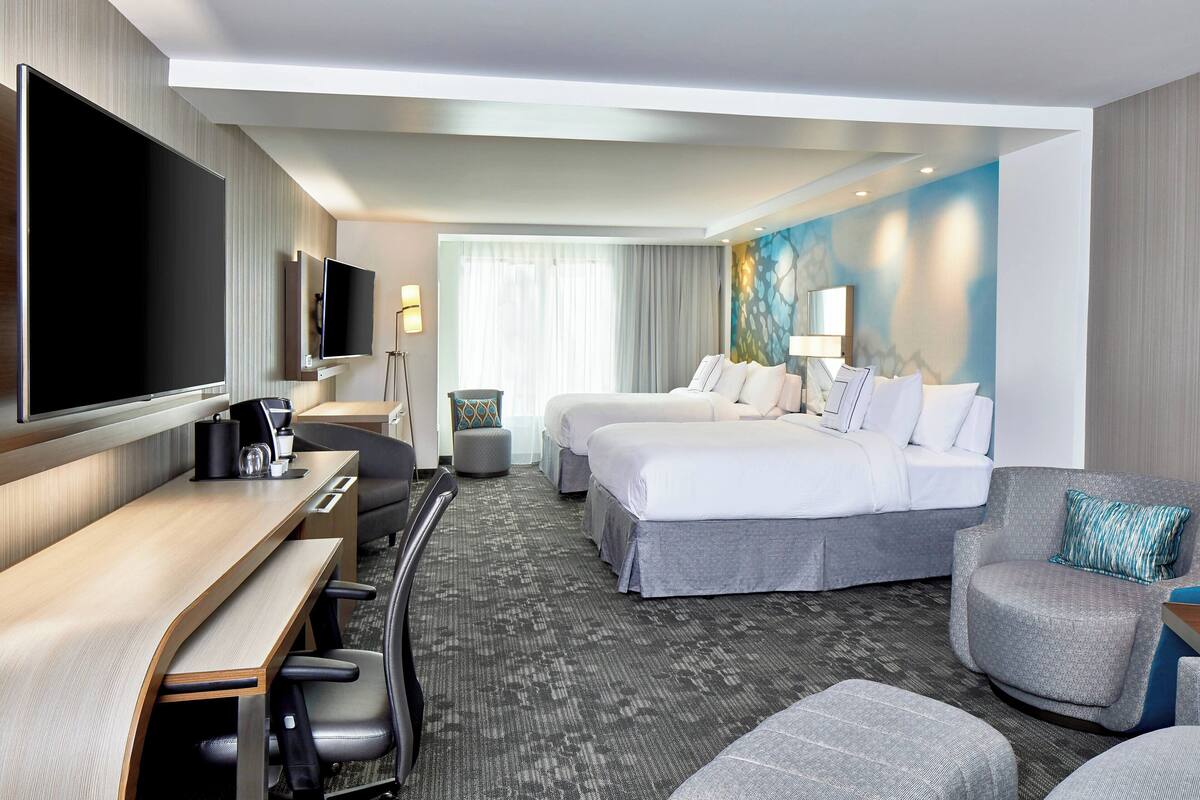 A hotel room with a large flat-screen TV, a desk with a chair, two beds, a sofa, and a small ottoman.