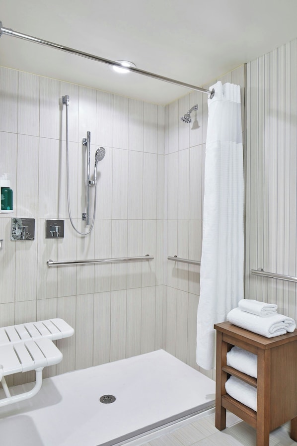 A modern bathroom with a walk-in shower, grab bars, a white bathtub, and a wooden side table with towels.