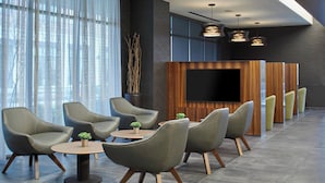 A modern hotel lobby with grey upholstered chairs, a round wooden table, and a flat-screen TV mounted on a wooden panel wall.