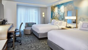 A hotel room with two beds, a desk, a chair, a lamp, and a large window with blinds.