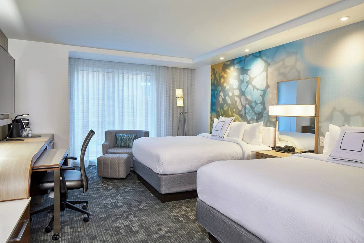 A hotel room with two beds, a desk, a chair, a lamp, and a large window with blinds.